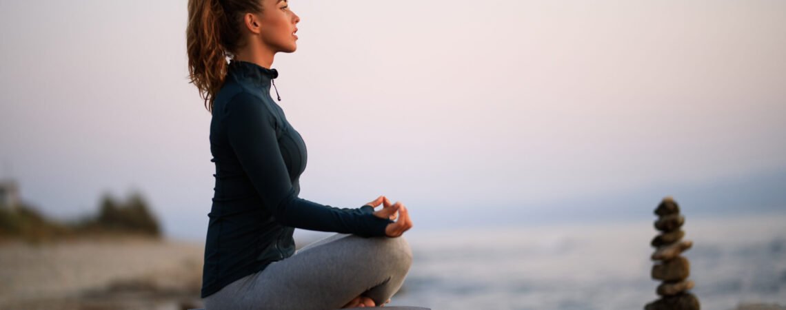 Athletic woman practicing Yoga and doing breathing exercise in lotus positing on a  beach rock.
